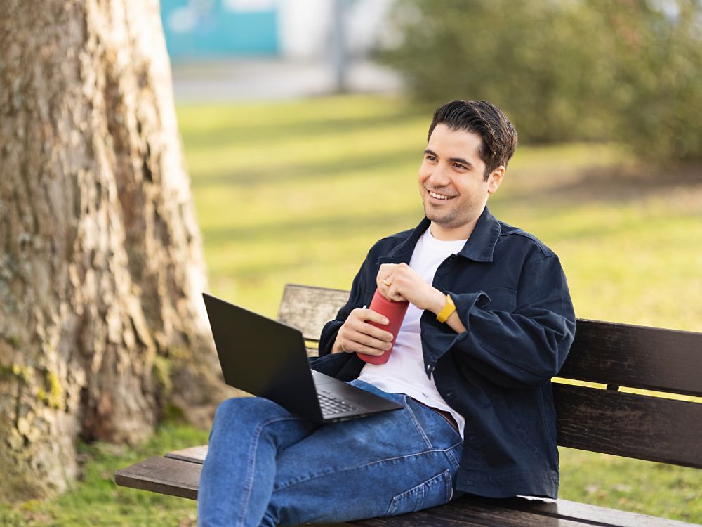 Pracownik Henkla siedzi na ławce w parku i pracuje na swoim laptopie. 