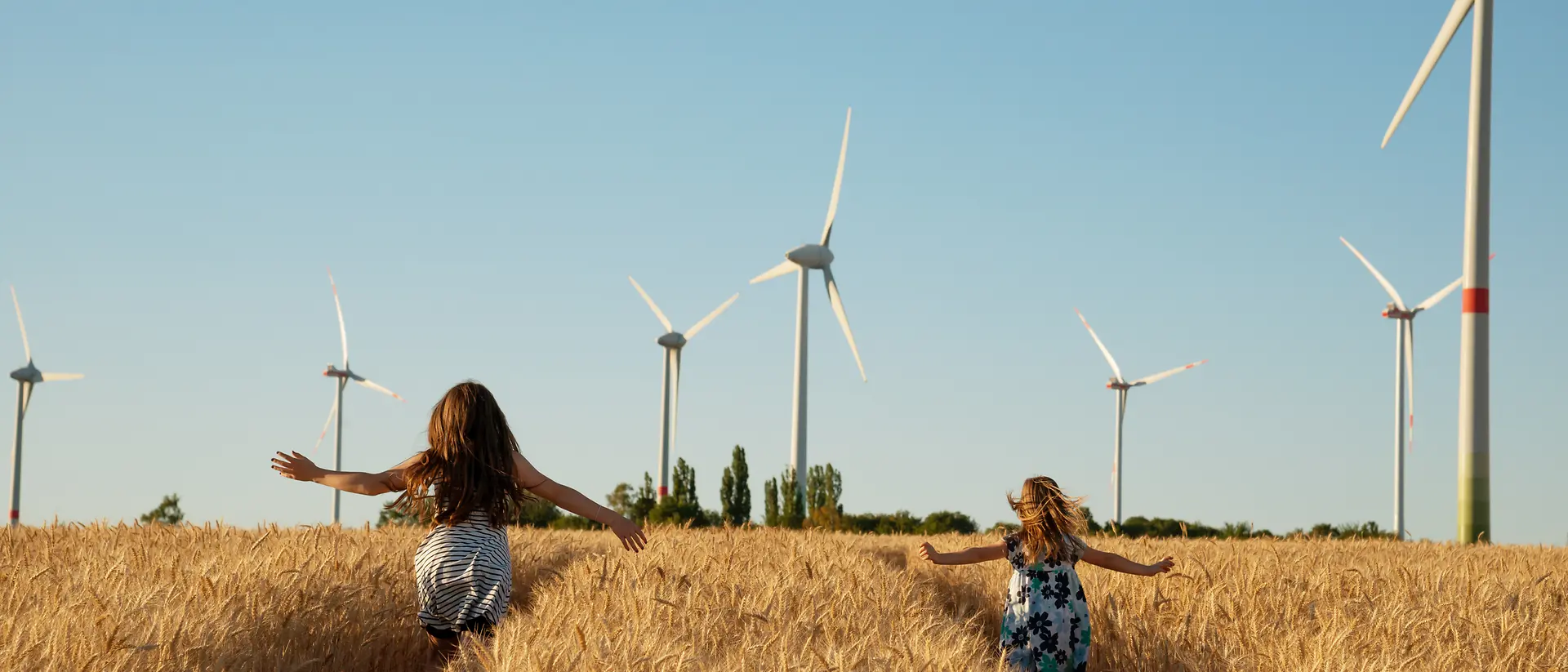 2 dziewczynki biegające po polu kukurydzy z elektrowniami wiatrowymi w tle