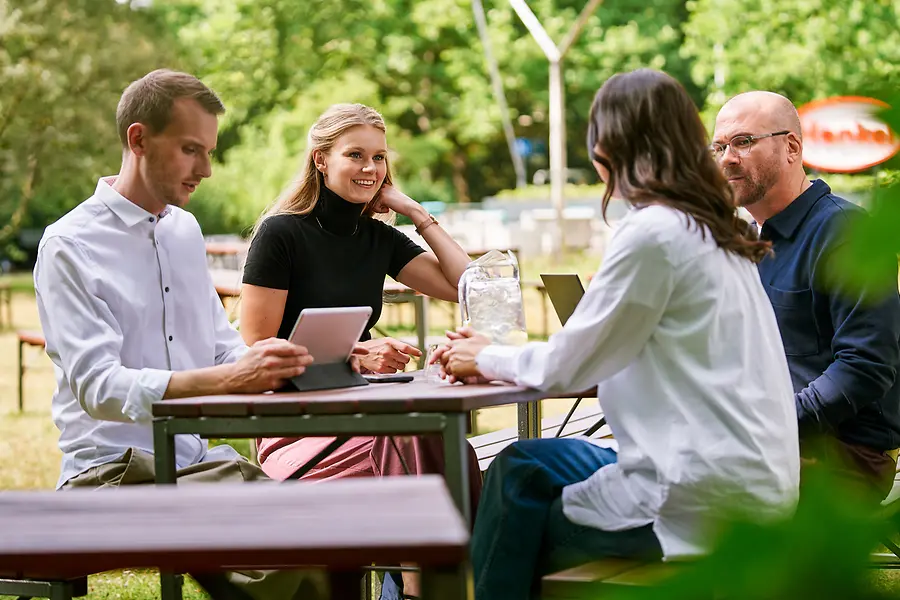 Grupa współpracowników siedzi razem na świeżym powietrzu, omawiając pracę przy pomocy tabletów i laptopów, co reprezentuje współpracę oraz otwartą, zorientowaną na zespół kulturę w Henkel.