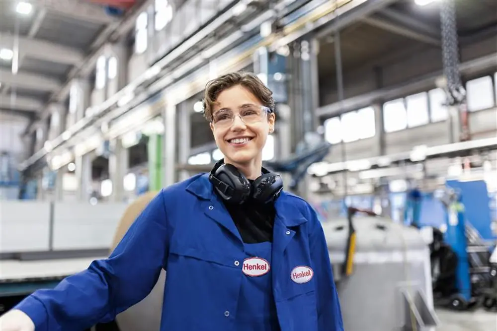 Uśmiechnięty pracownik Henkel w niebieskim płaszczu roboczym i słuchawkach ochronnych, stojący w hali produkcyjnej, reprezentujący wiedzę i innowacje w produkcji.