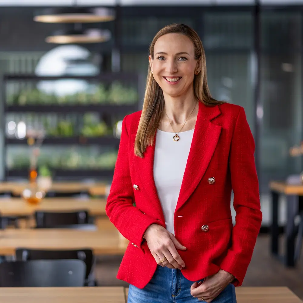 Dr Simone Bagel-Trah w jasnoczerwonej marynarce i białej bluzce, stojąca w nowoczesnym wnętrzu w stylu kawiarni z drewnianymi stolikami i dekoracyjnymi wazonami w tle.