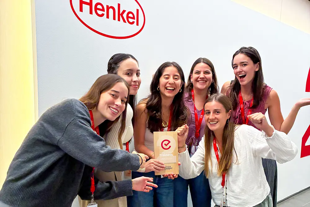 Grupa sześciu osób stoi razem przed ścianą z logo Henkel. Mają na sobie czerwone smycze z identyfikatorami i swobodne stroje i stoją blisko siebie wokół tabliczki z nagrodą. Scena to wewnętrzna przestrzeń korporacyjna z jasną ścianą i szarą podłogą. Grupa entuzjastycznie pozuje, podkreślając nagrodę znajdującą się pośrodku.
