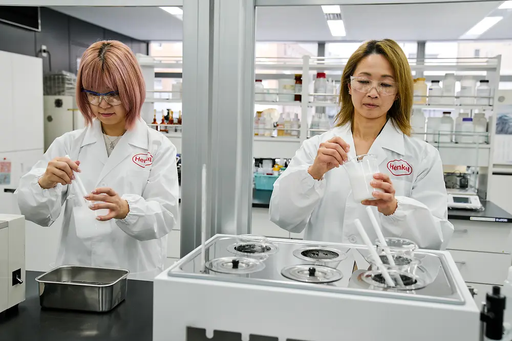Dwie osoby w białych fartuchach laboratoryjnych z logo Henkel pracują w nowoczesnym laboratorium. Obsługują przyrządy naukowe i pojemniki na blacie, a w tle widać różne urządzenia laboratoryjne i półki z materiałami eksploatacyjnymi.