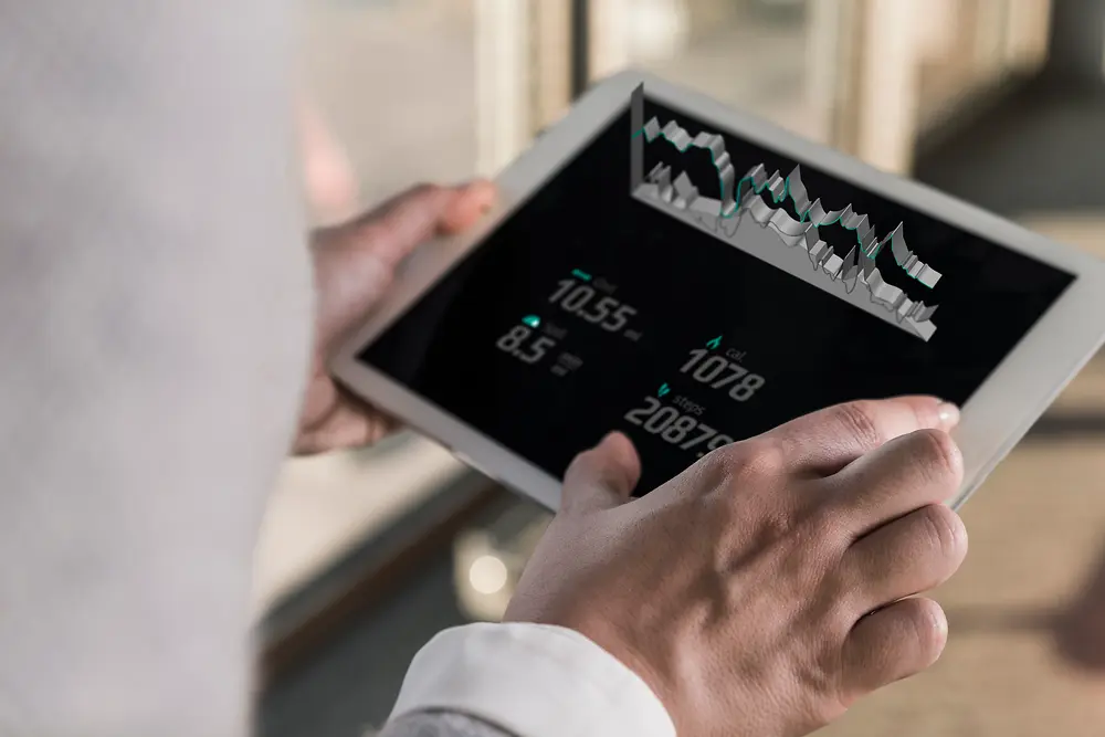 Osoba trzymająca tablet z wyświetlanymi wykresami cyfrowymi i danymi liczbowymi, widziana z perspektywy osoby stojącej za nią w jasnym pomieszczeniu.