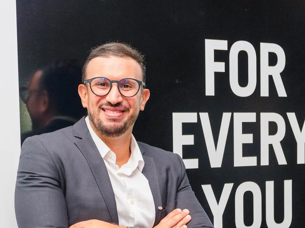 Employee in business attire standing with arms crossed in front of ‘FOR EVERY YOU’ branded backdrop