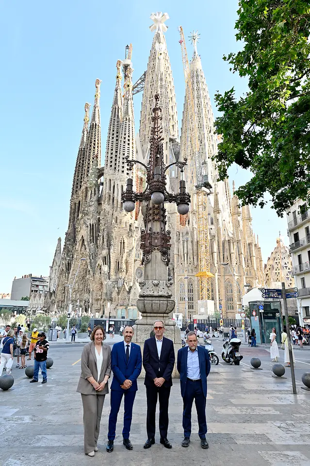 Vista general de la Sagrada Familia en Barcelona con sus torres centrales en construcción y cuatro personas posando en primer plano frente al templo en una plaza urbana con peatones y elementos de mobiliario urbano.