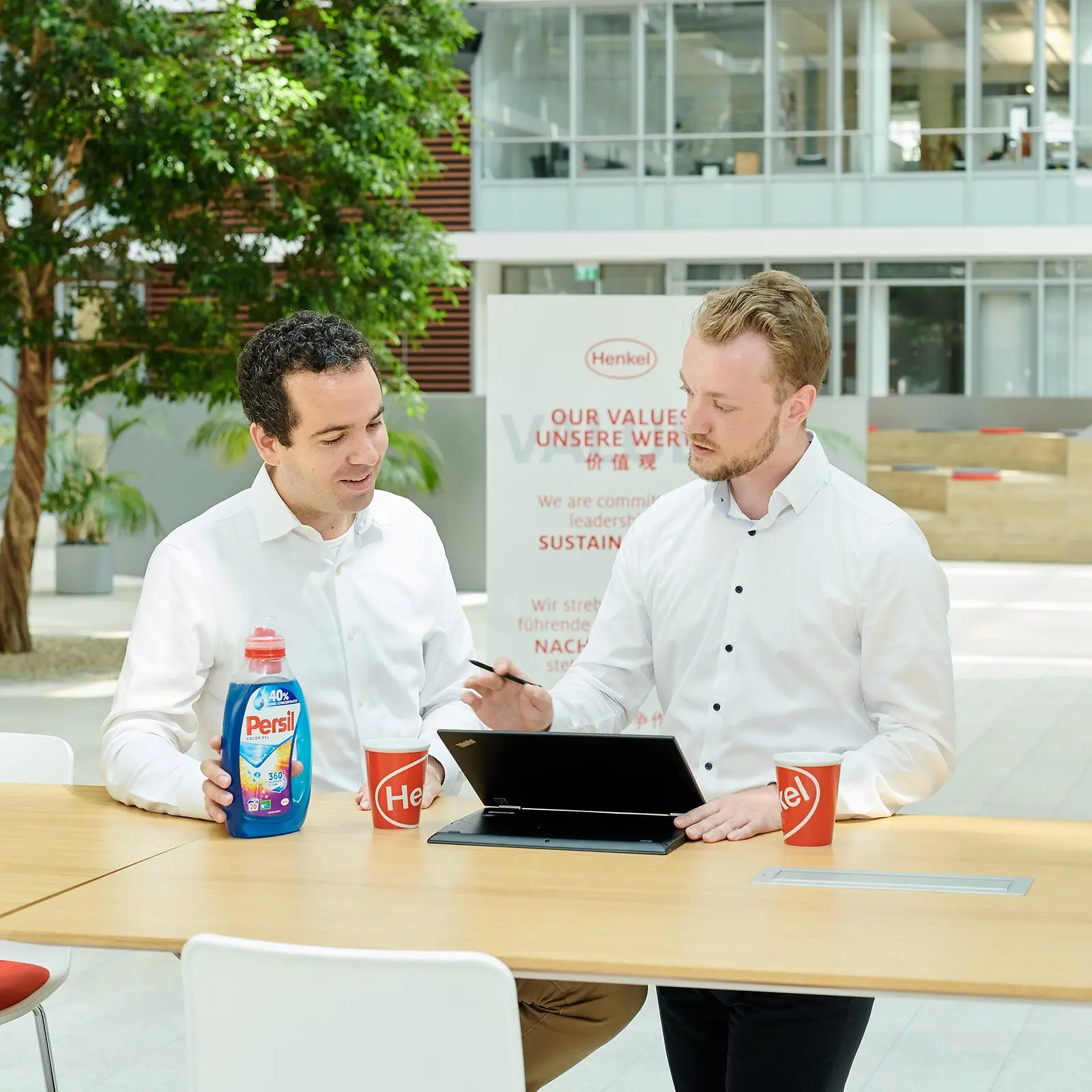 Dwóch mężczyzn stojących przy stole z butelką Persil i laptopem